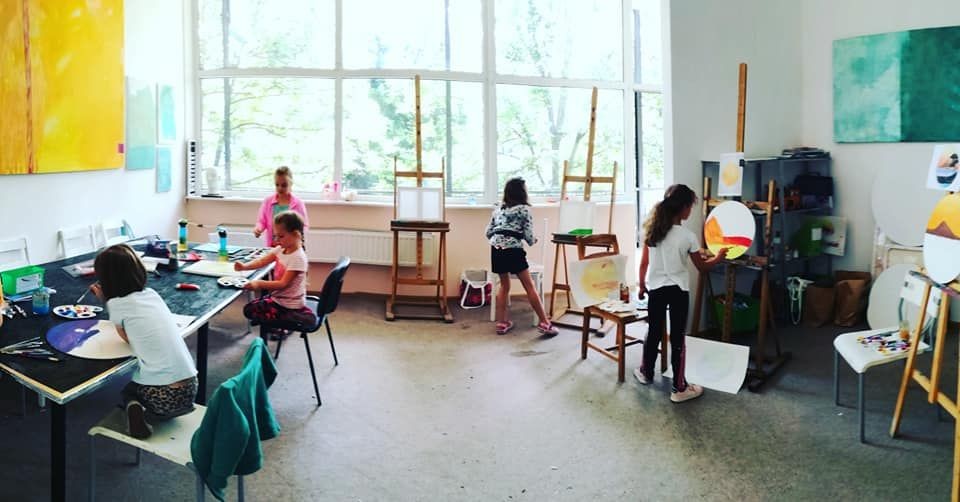 Children painting on canvases in a sunlit art classroom with easels and art supplies around.