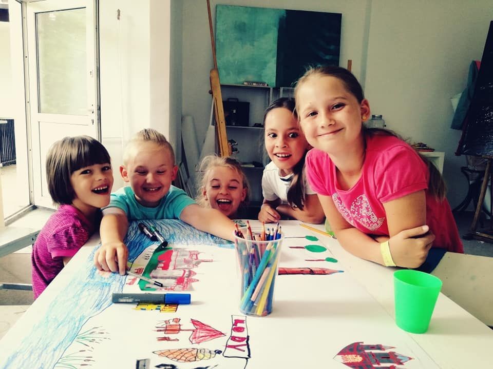 Five children smiling and leaning over a table filled with drawings and colored pencils in an art classroom.