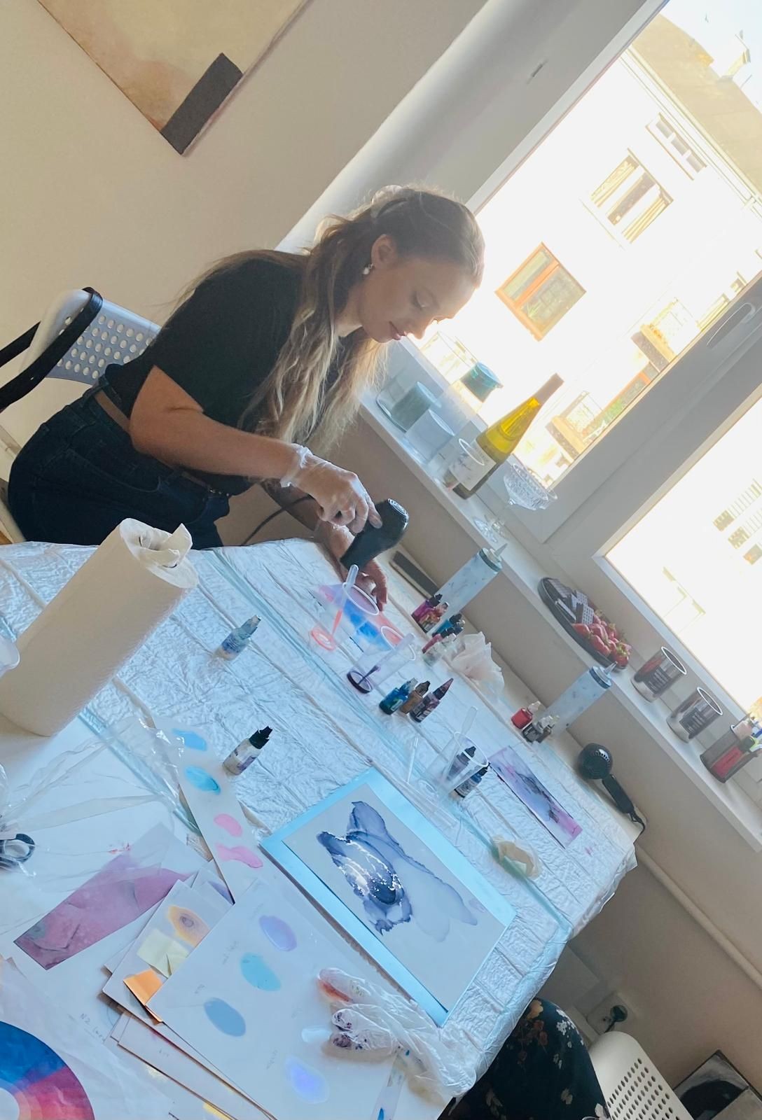 Woman sitting at a table near a window, working with art supplies and creating abstract paintings.