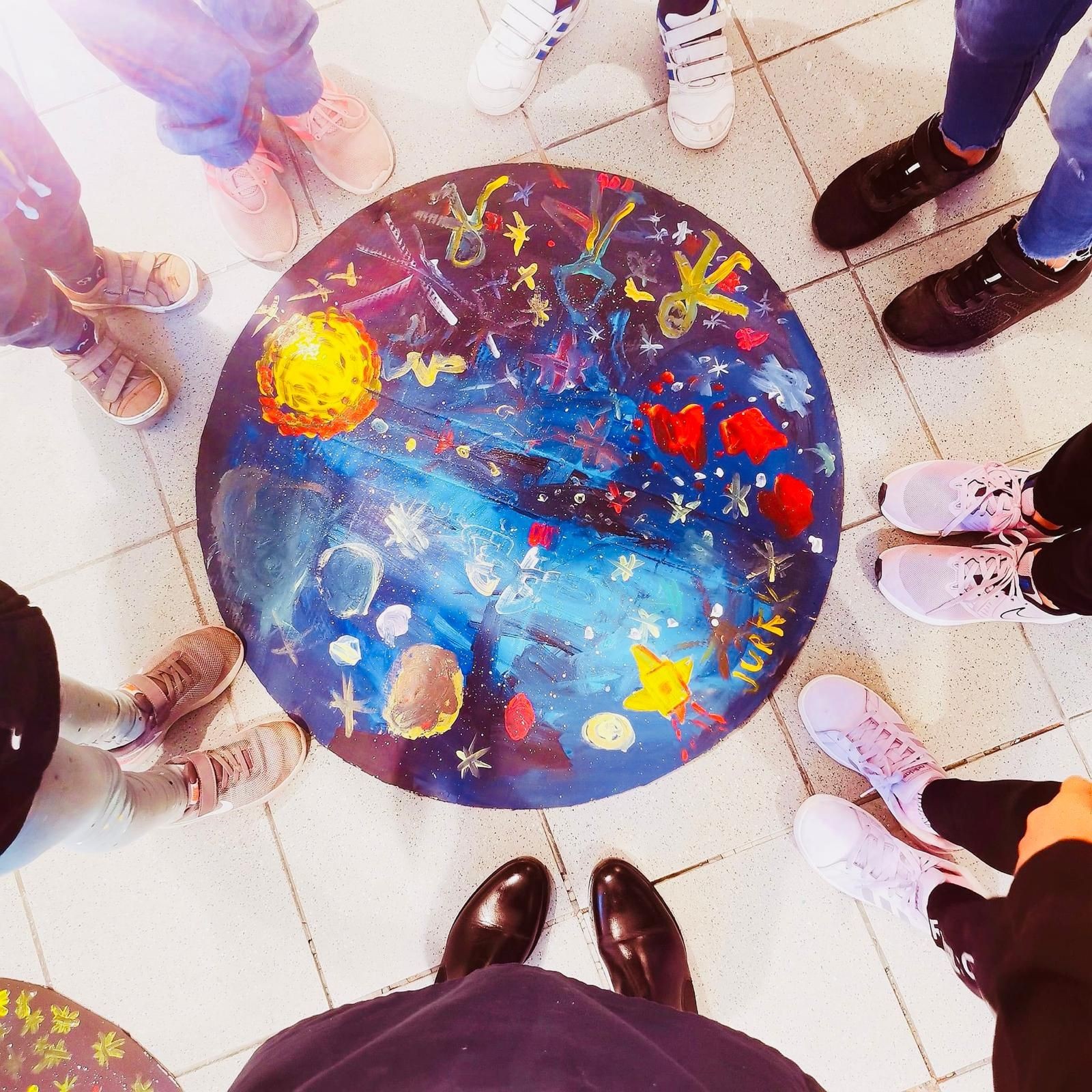 Group of people standing in a circle around a colorful galaxy-themed painting on the floor.